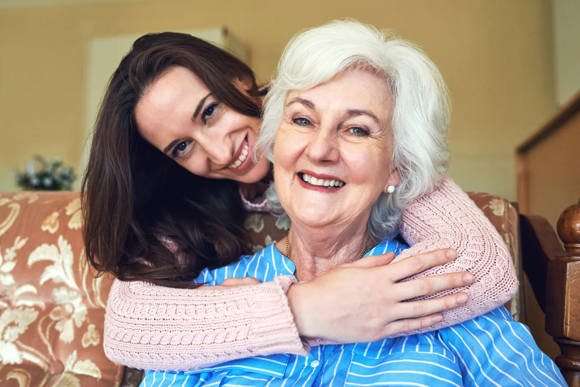 Young woman stands behind a seated senior living resident and hugs her around the neck as they both smile