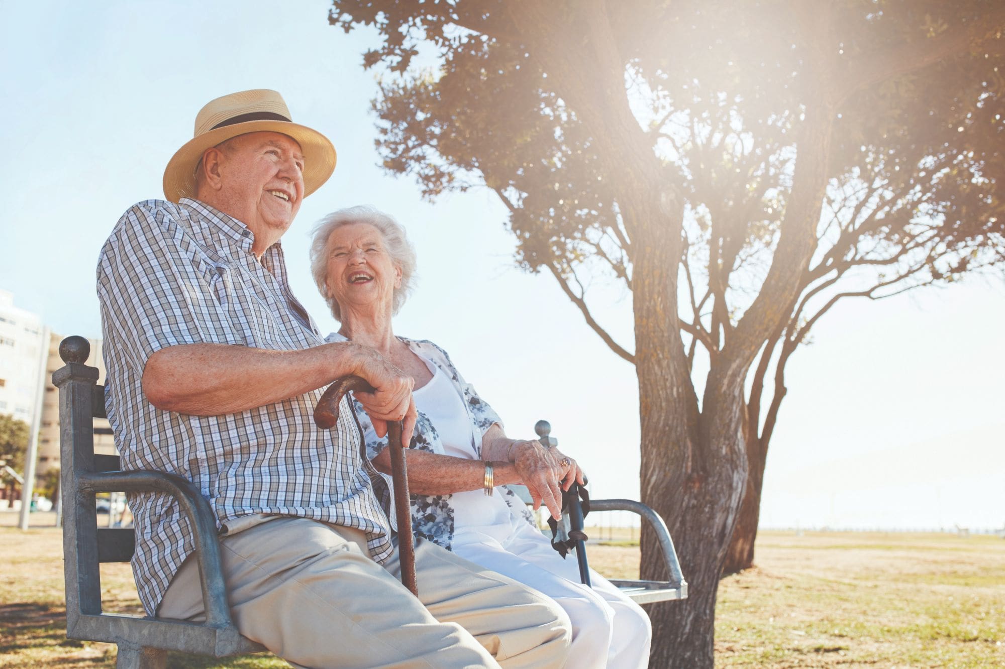 Male and female Flourish Senior Living residents smile and sit together on an outdoor bench next to a tree Male and female Flourish Senior Living residents smile and sit together on an outdoor bench next to a tree