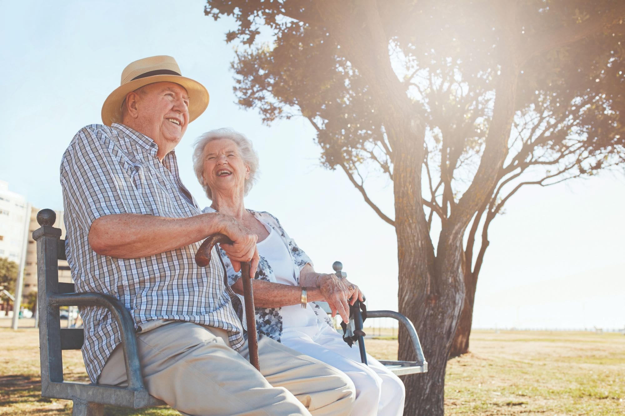 Male and female Flourish Senior Living residents smile and sit together on an outdoor bench next to a tree