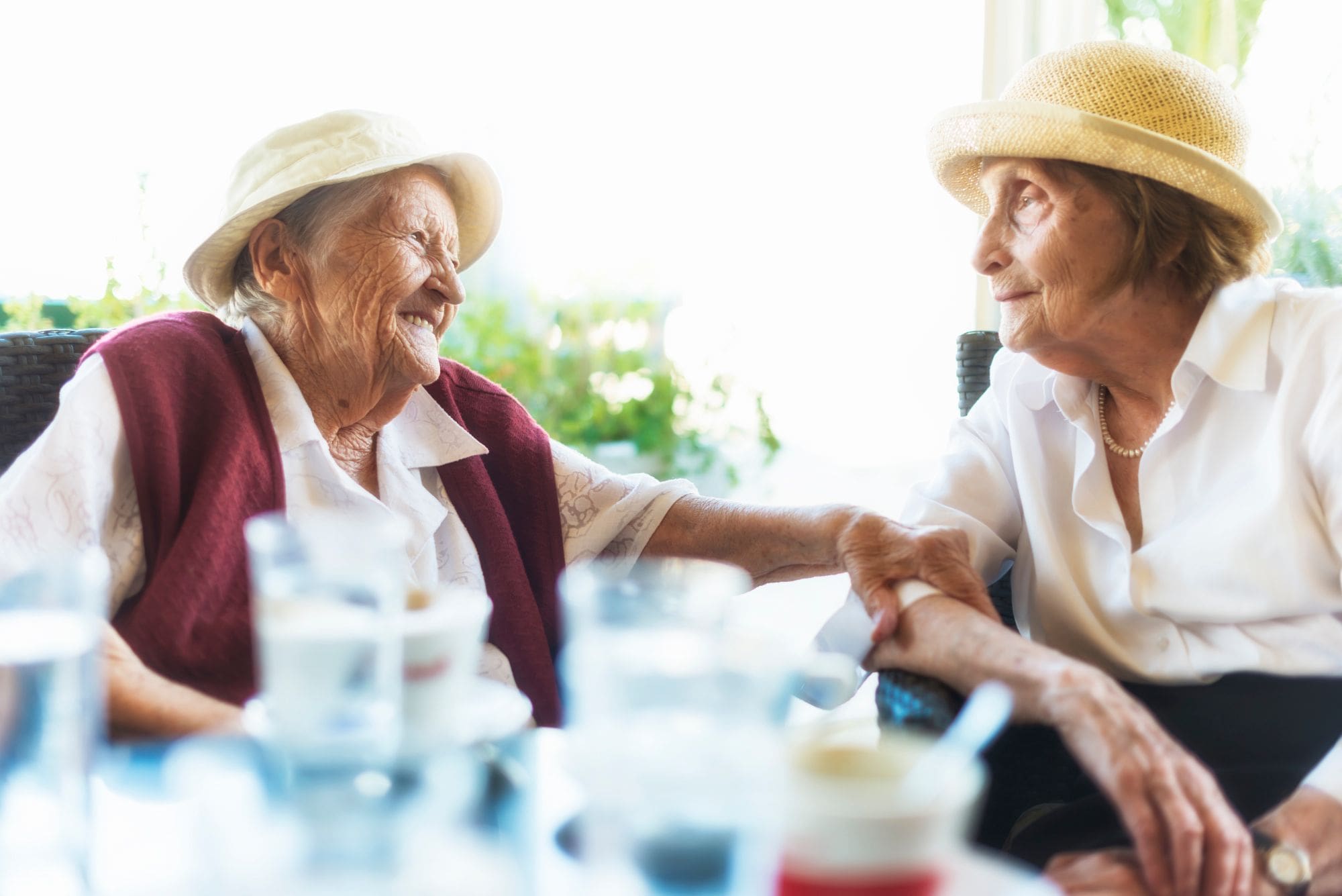 Senior Living residents holding the arm of another older woman as they sit wearing hats at an outdoor dining table and smile Senior Living residents holding the arm of another older woman as they sit wearing hats at an outdoor dining table and smile