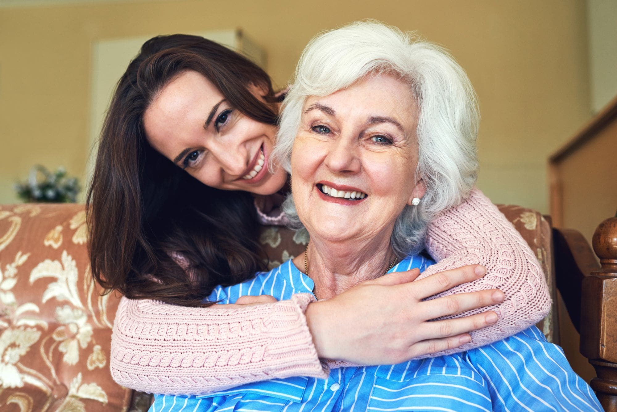 Young woman stands behind a seated senior living resident and hugs her around the neck as they both smile