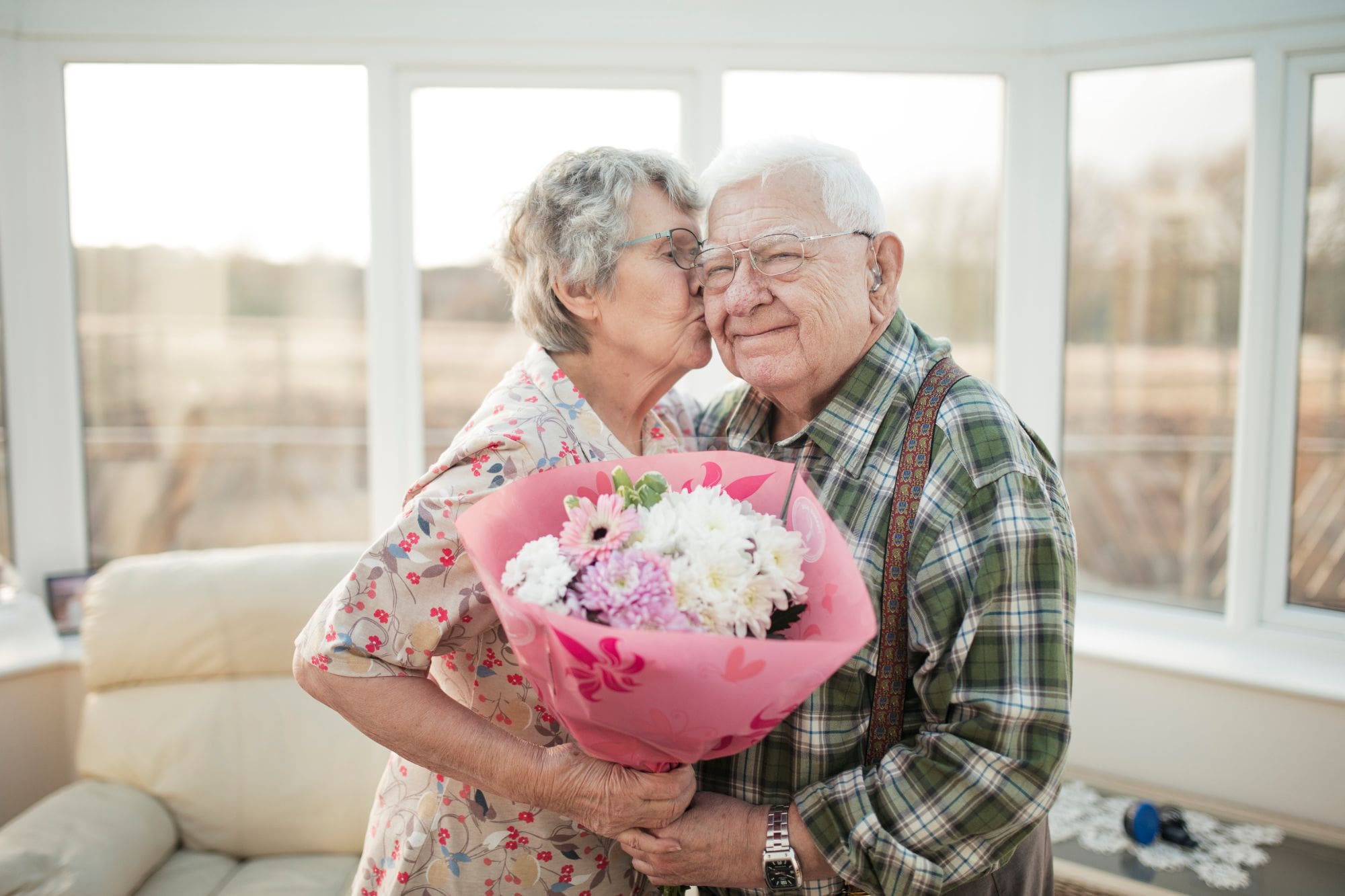 Senior aged female gives a kiss on the cheek to older male while they hold a flower bouquet in front of a large window